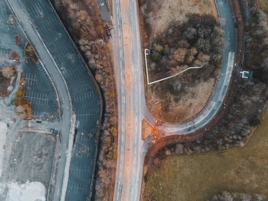 An aerial view showing a multi-lane road running vertically through the center of the image, with white markings delineating lanes and directional arrows. To the left, there is a large, mostly empty p