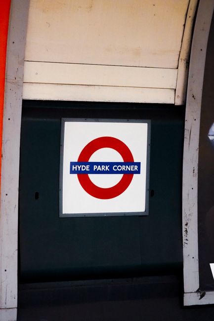Close-up of a square sign mounted on a dark green structure, displaying the London Underground roundel with a white background, a red circle, and a blue horizontal bar with white text reading 'HYDE PA